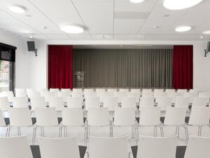 Bild vergrößern: weiße Stuhlreihen und im Hintergrund die Bühne mit rotem Vorhang