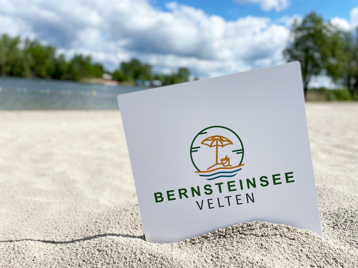 Bild vergrößern: Sonnenschirm und Buddeleimer  - Das neue Logo des Veltener Bernsteinsees seit 2022.